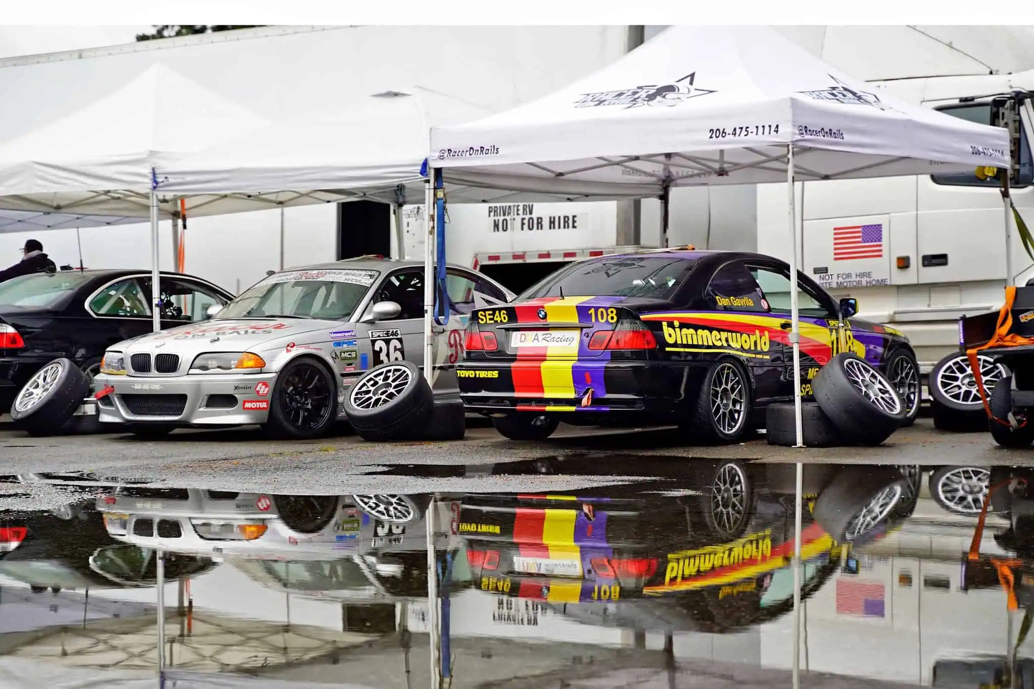 Racer on Rails-supported Spec E46 BMW race cars beneath paddock tents, reflected in rainwater at Pacific Raceways.