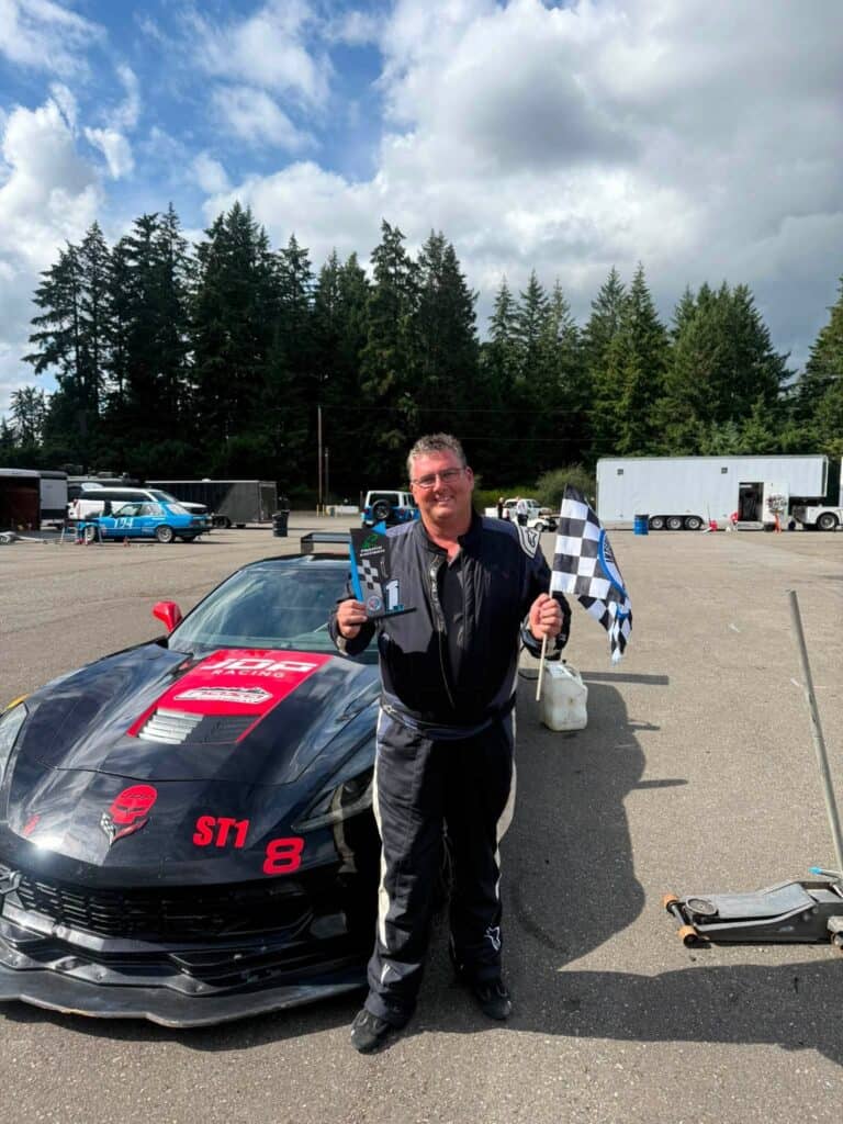 Jay Perry holds a first-place trophy and checkered flag beside his black C7 Corvette at Pacific Raceways.